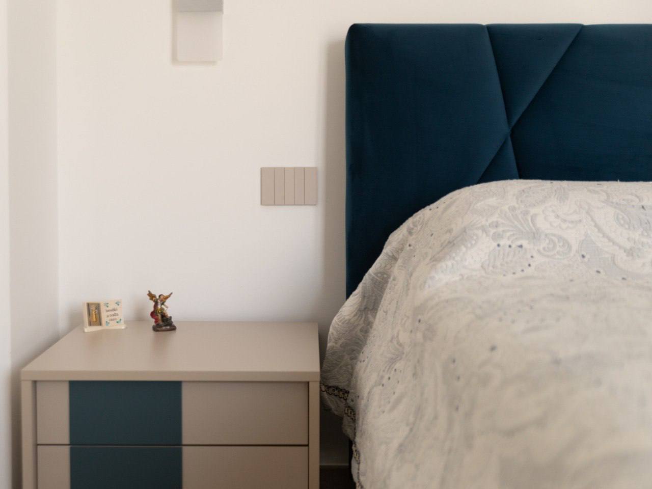 Dettaglio di comodino tortora e blu accanto alla testiera imbottita in velluto